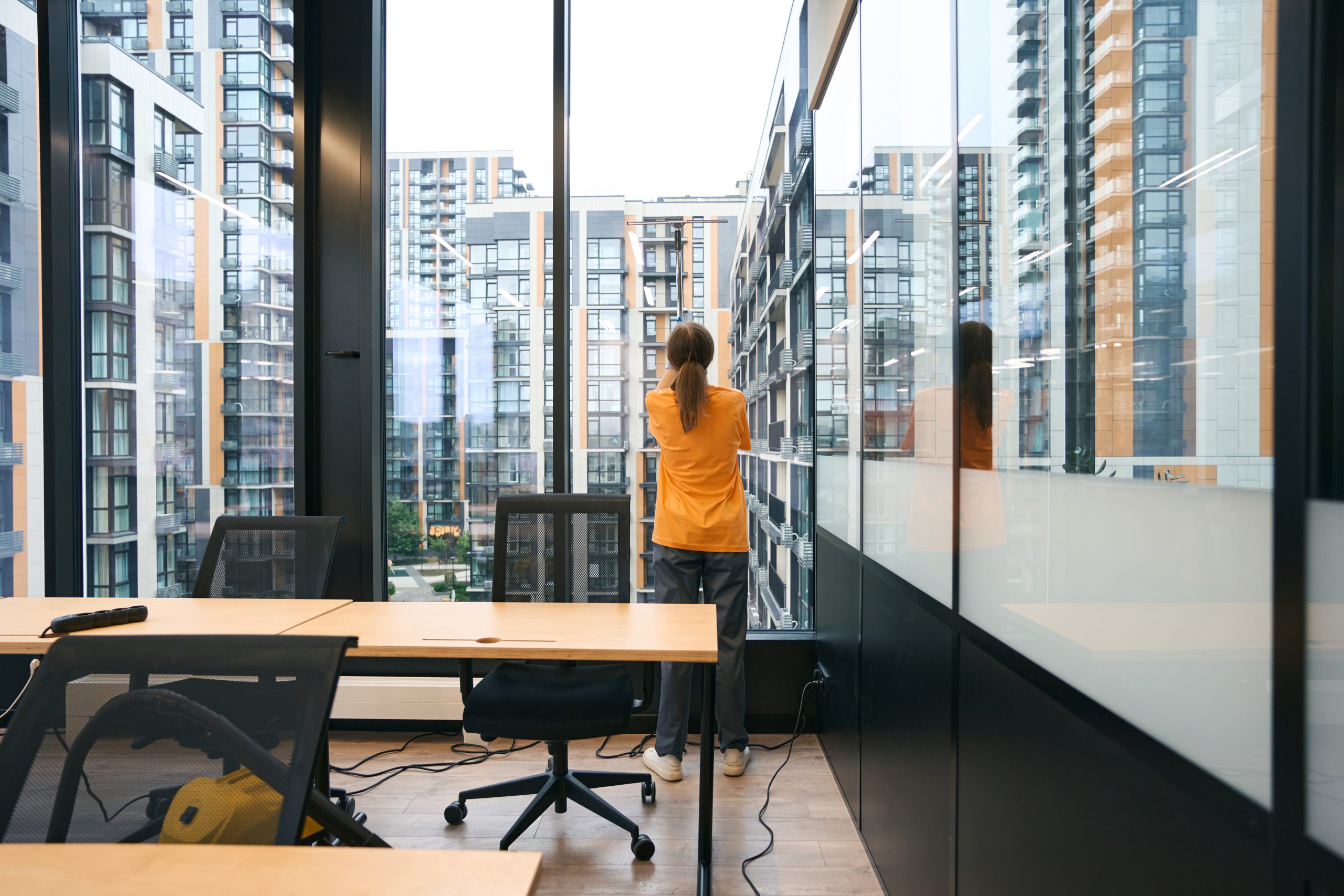 Employee of the cleaning service is resting and looking out the window, she is wearing yellow overalls, next to a vacuum cleaner